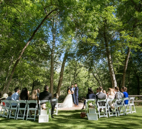 A wedding ceremony on a green lawn, with guests seated in white chairs facing the couple under a canopy of leafy trees.