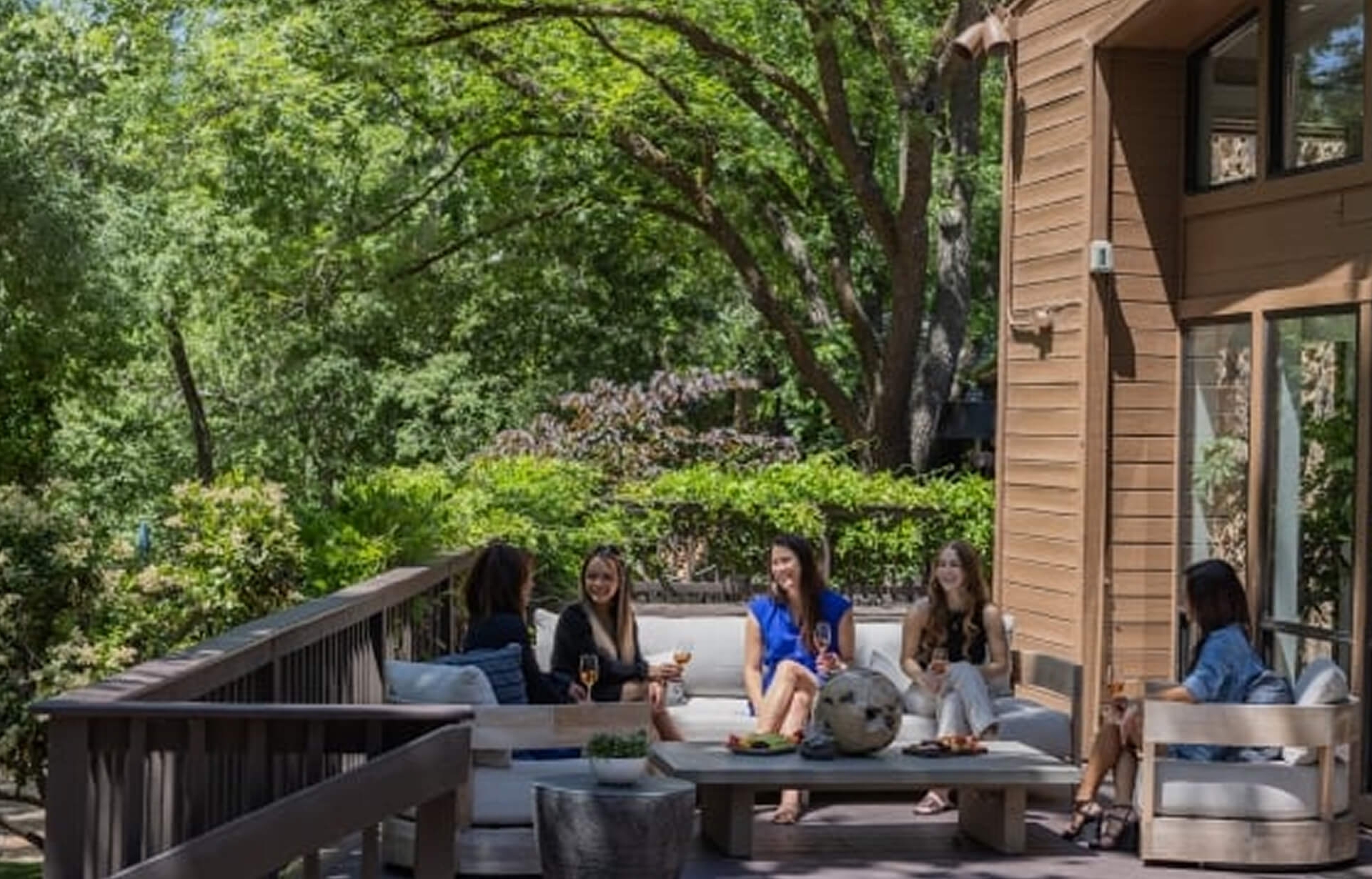 A group of women relaxing with drinks on a comfortable outdoor deck at the Creekhouse, shaded by large trees.
