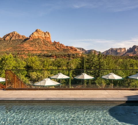 View from a swimming pool, looking past white umbrellas on the deck to a clear, panoramic view of Sedona's red rocks.