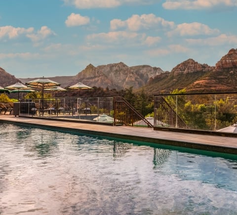 View from the edge of a resort pool, looking over the water at the deck, umbrellas, and a panorama of Sedona's red rocks.