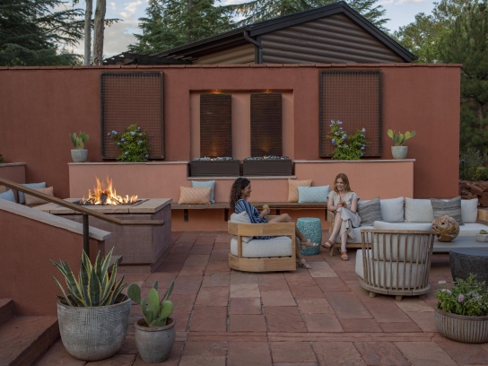 Two women talk while sitting in comfortable chairs on a red-tiled patio, gathered around a large, modern fire pit.