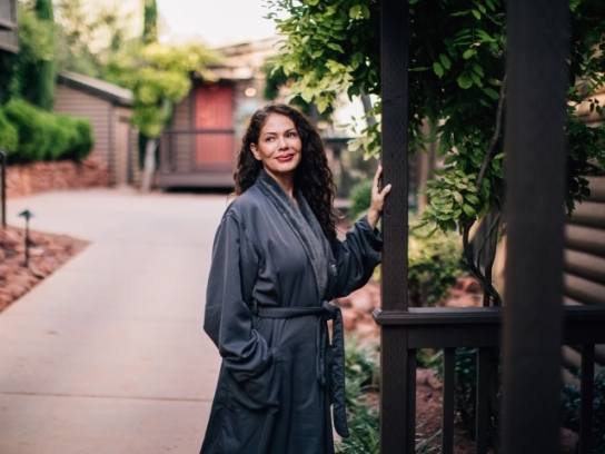 A smiling woman with long curly hair, wearing a gray robe, stands on a garden path next to a log cabin.