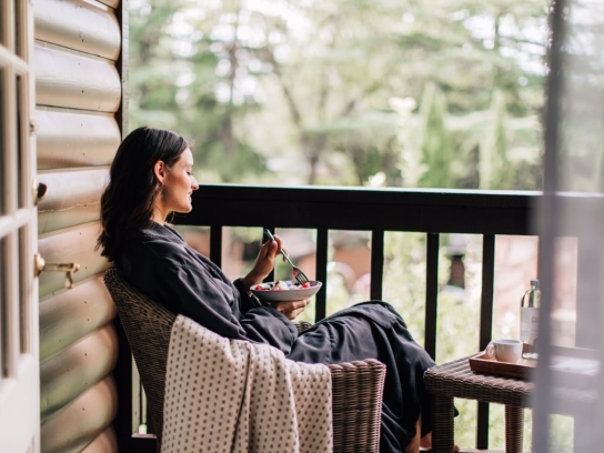 A woman in a robe relaxes with a bowl of fruit on a wicker chair on a private, wooded cottage balcony.