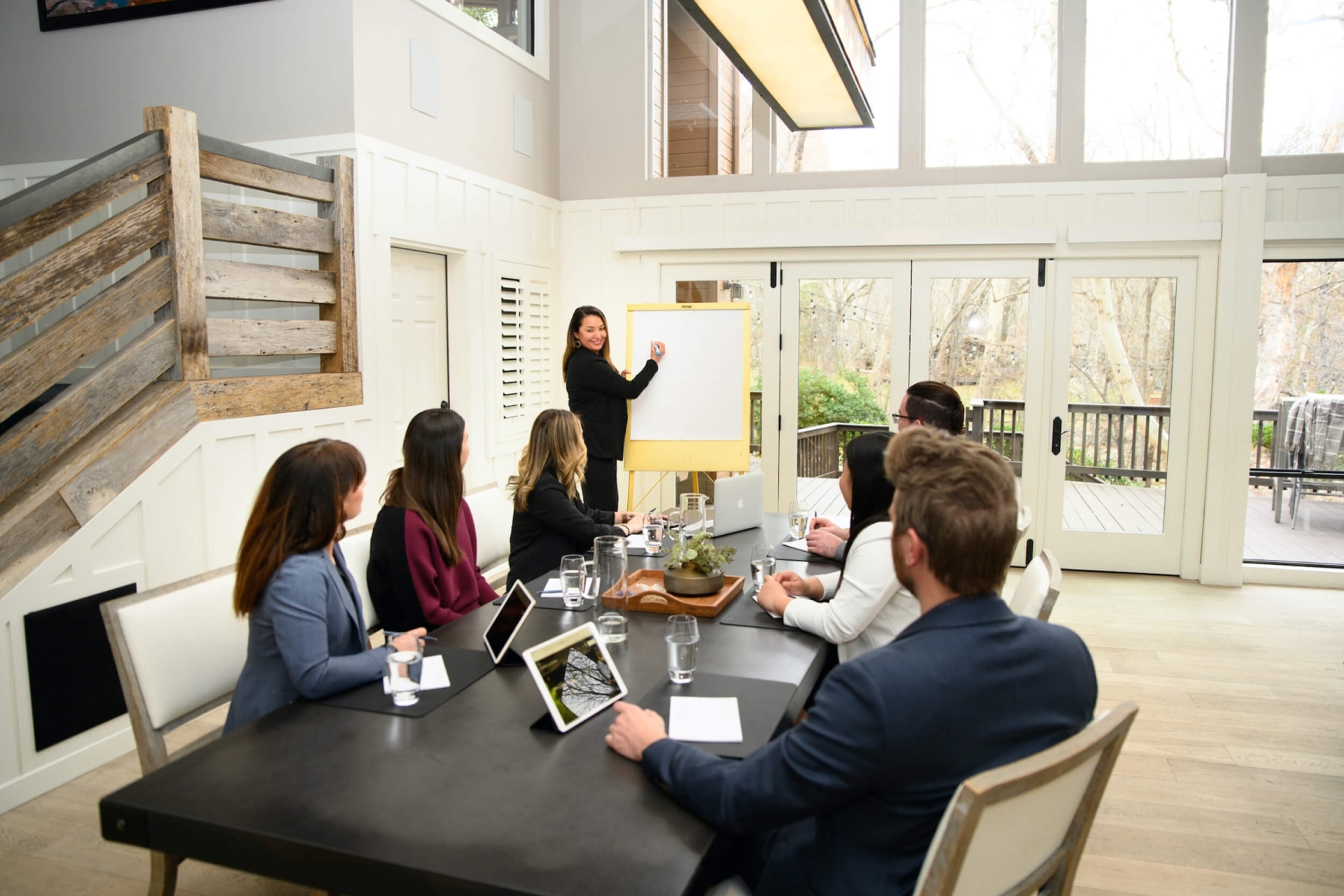 A woman presents on a whiteboard to a group of colleagues seated at a long table in a bright, modern meeting room.