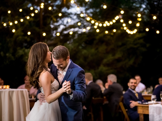 A bride and groom smile and embrace during their first dance at an outdoor wedding reception under string lights.