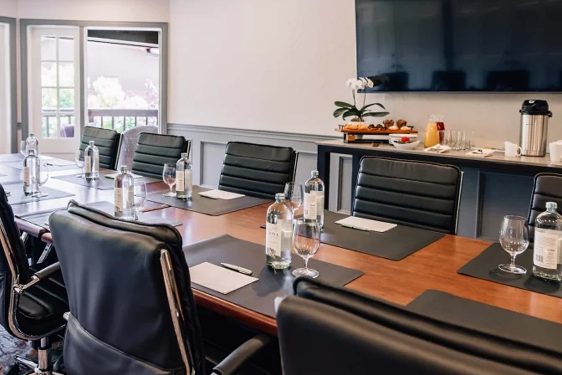 A professional boardroom with a long wooden table, black leather chairs, a large screen, and a refreshment station.
