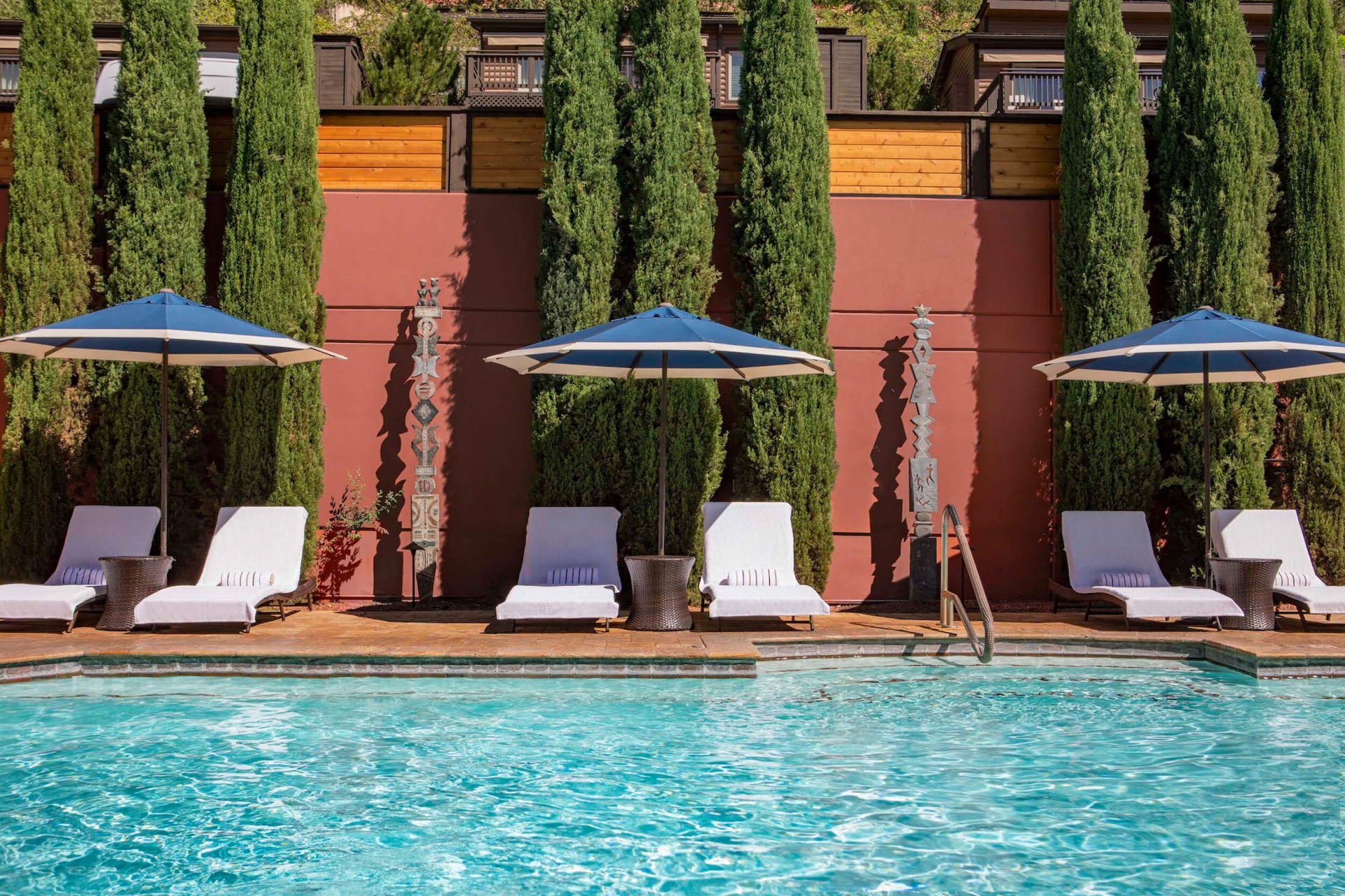 White lounge chairs and blue-striped umbrellas line a sparkling blue pool, set against a red wall and tall cypress trees.