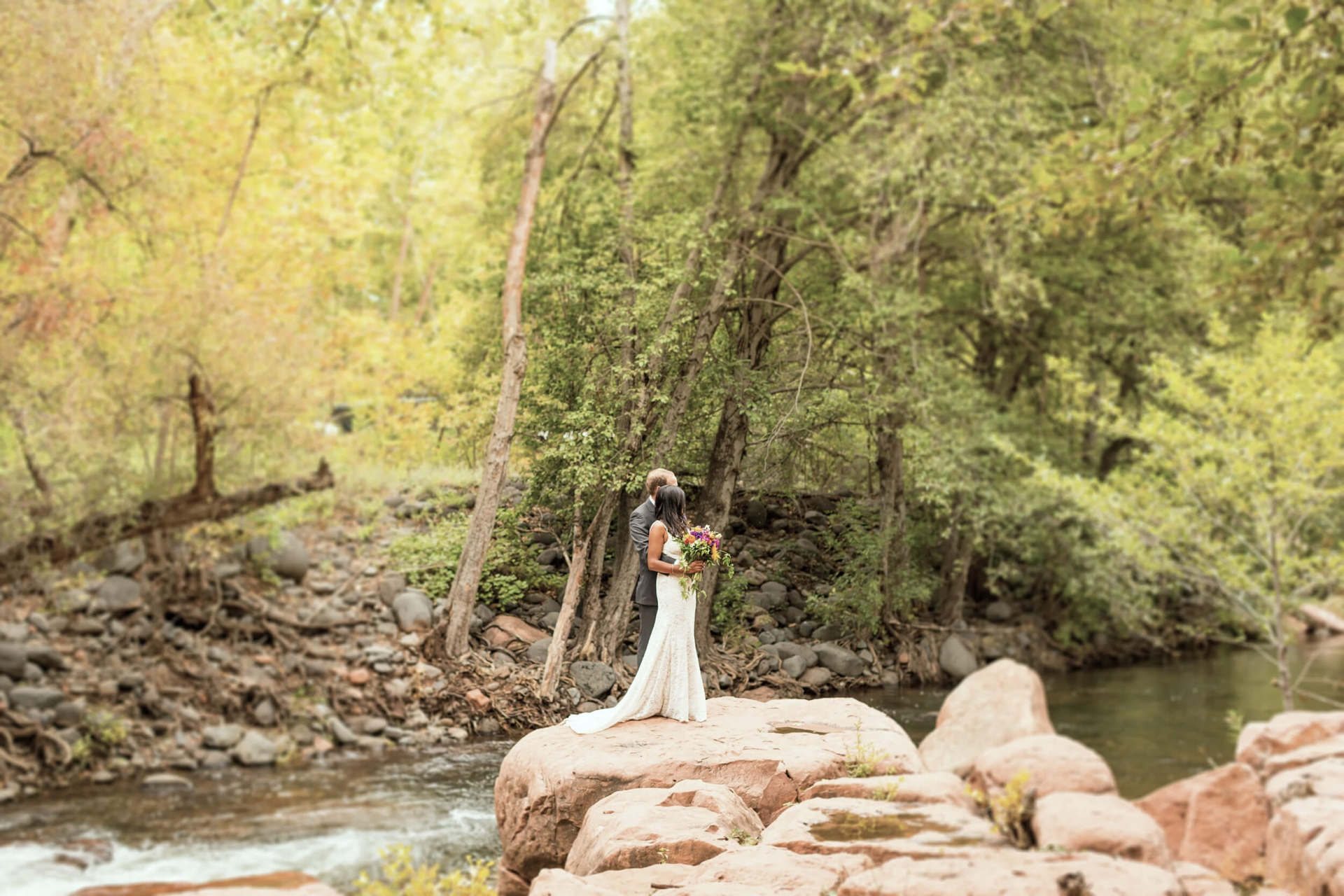 A bride and groom embrace on a large rock in the middle of Oak Creek, surrounded by lush green trees.