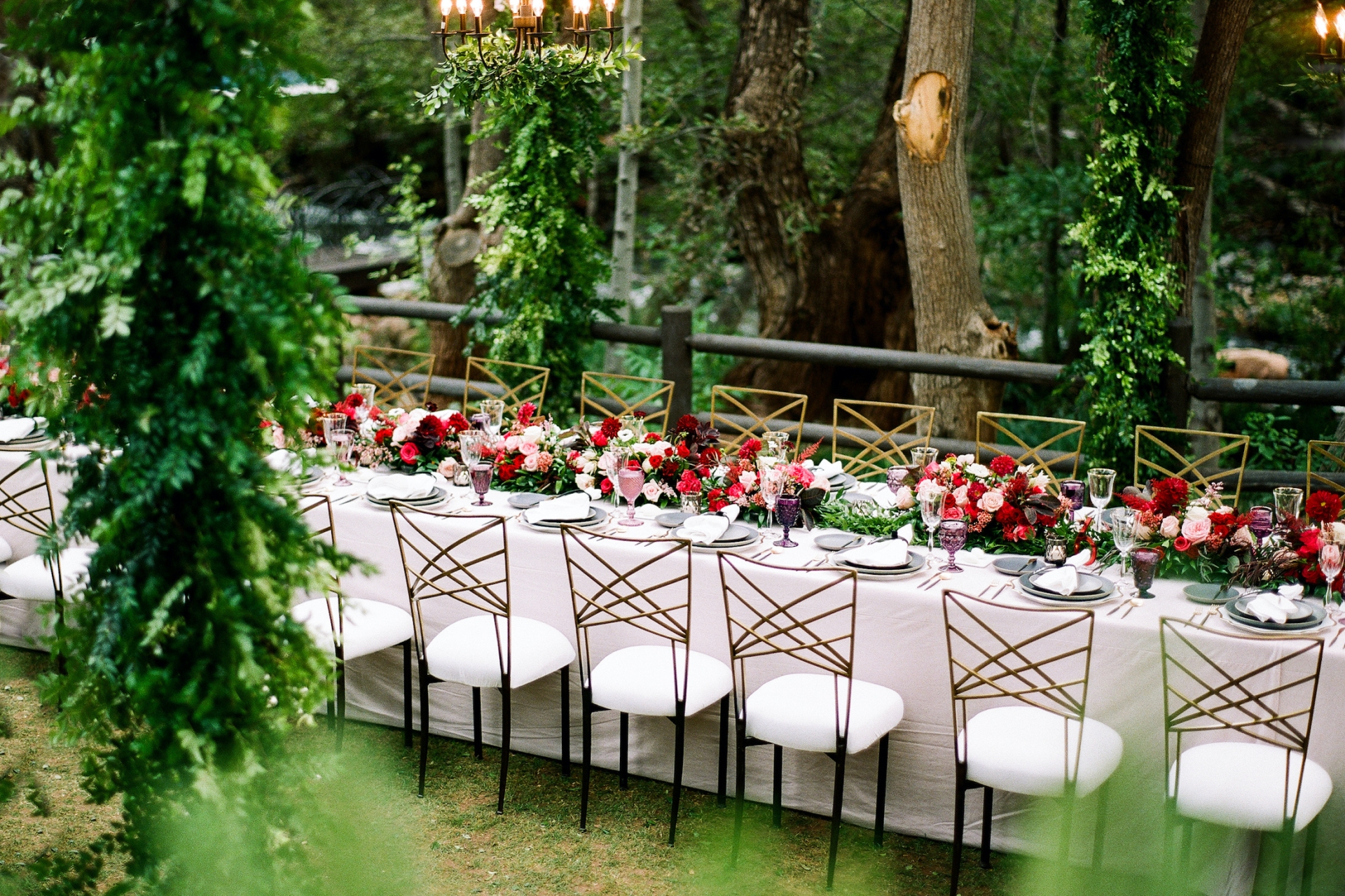 An elegant, long wedding table set on a lawn, decorated with red floral runners and lined with gold and white chairs.