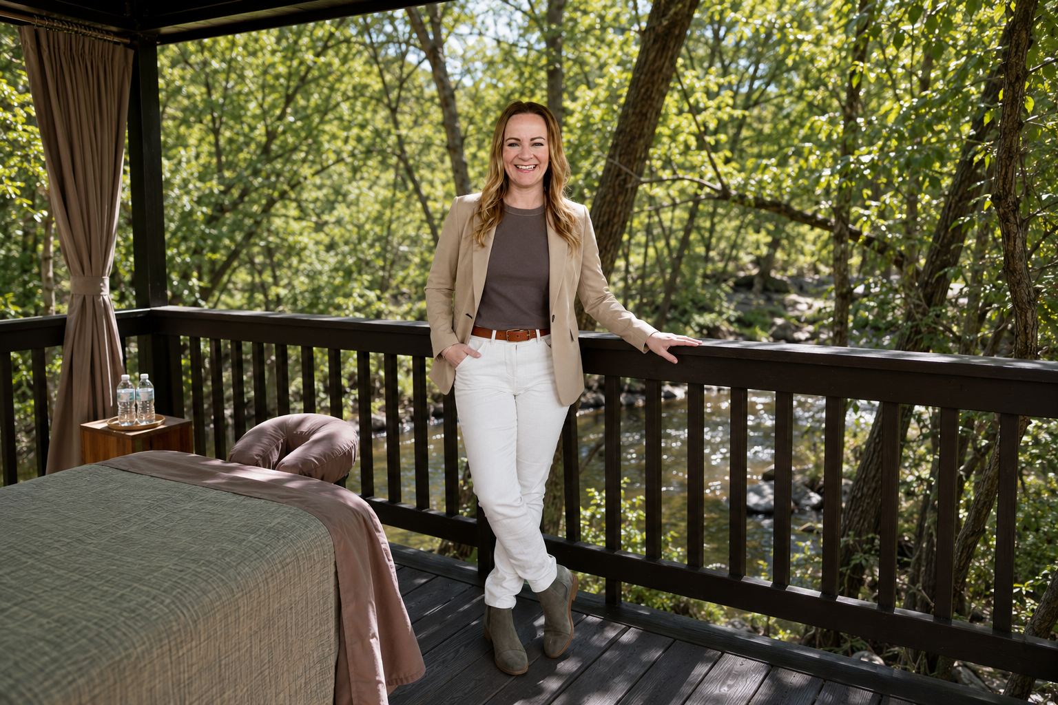 Smiling woman stands on a wooden deck in a forest by a river, wearing a beige blazer, grey top, white pants, brown belt, and grey ankle boots.